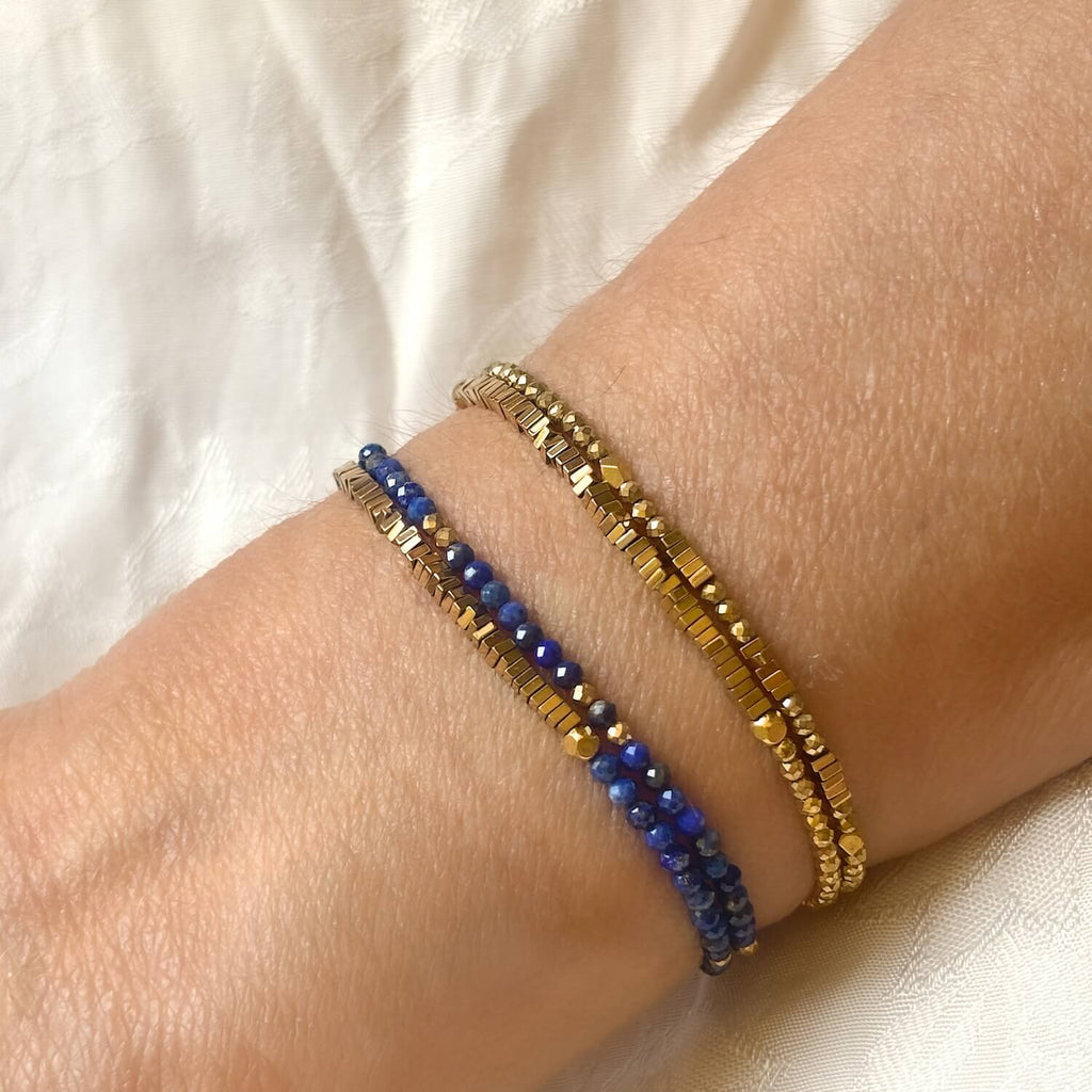 Two bracelets, one gold and one blue,  golden Hematite and Lapis Lazuli wrap double strand gemstone bracelets on a wrist against a neutral background. 