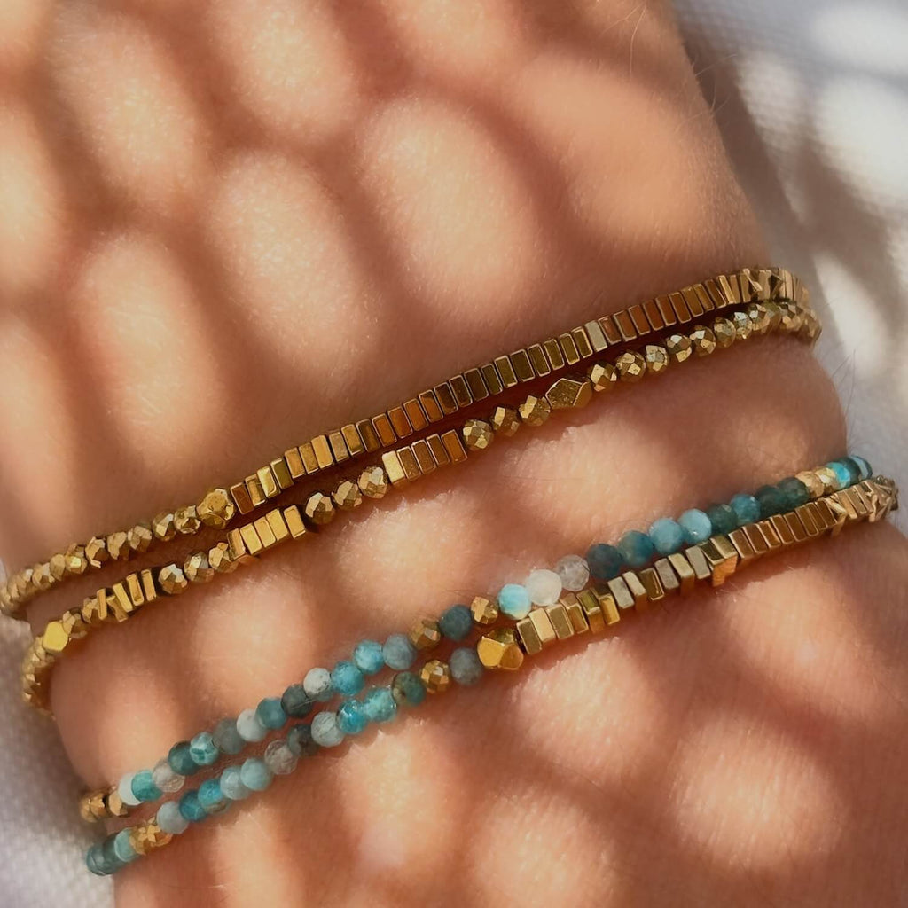 Close-up of a wrist wearing 2 beaded gemstone bracelets with golden Hematite and Blue Apatite gemstones  against a white fabric sunlit background with artistic shadows