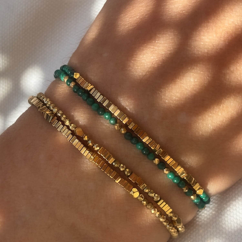 Two bracelets, one gold and one green,  golden Hematite and Malachite wrap beaded bracelets on a wrist against a white fabric sunlit background. 