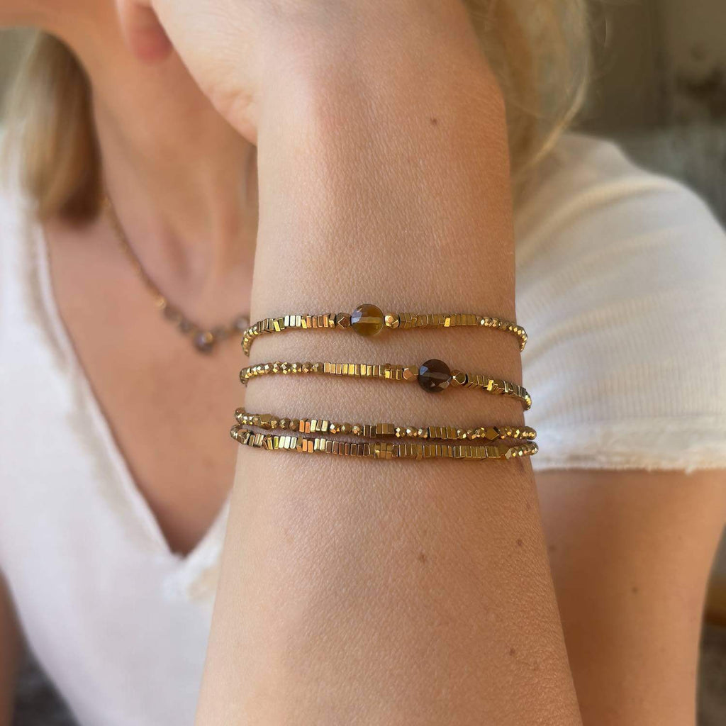 Close-up of a person's wrist wearing multiple gold en hematite , citrine and smoky quartz gemstone bracelets on a blurred background