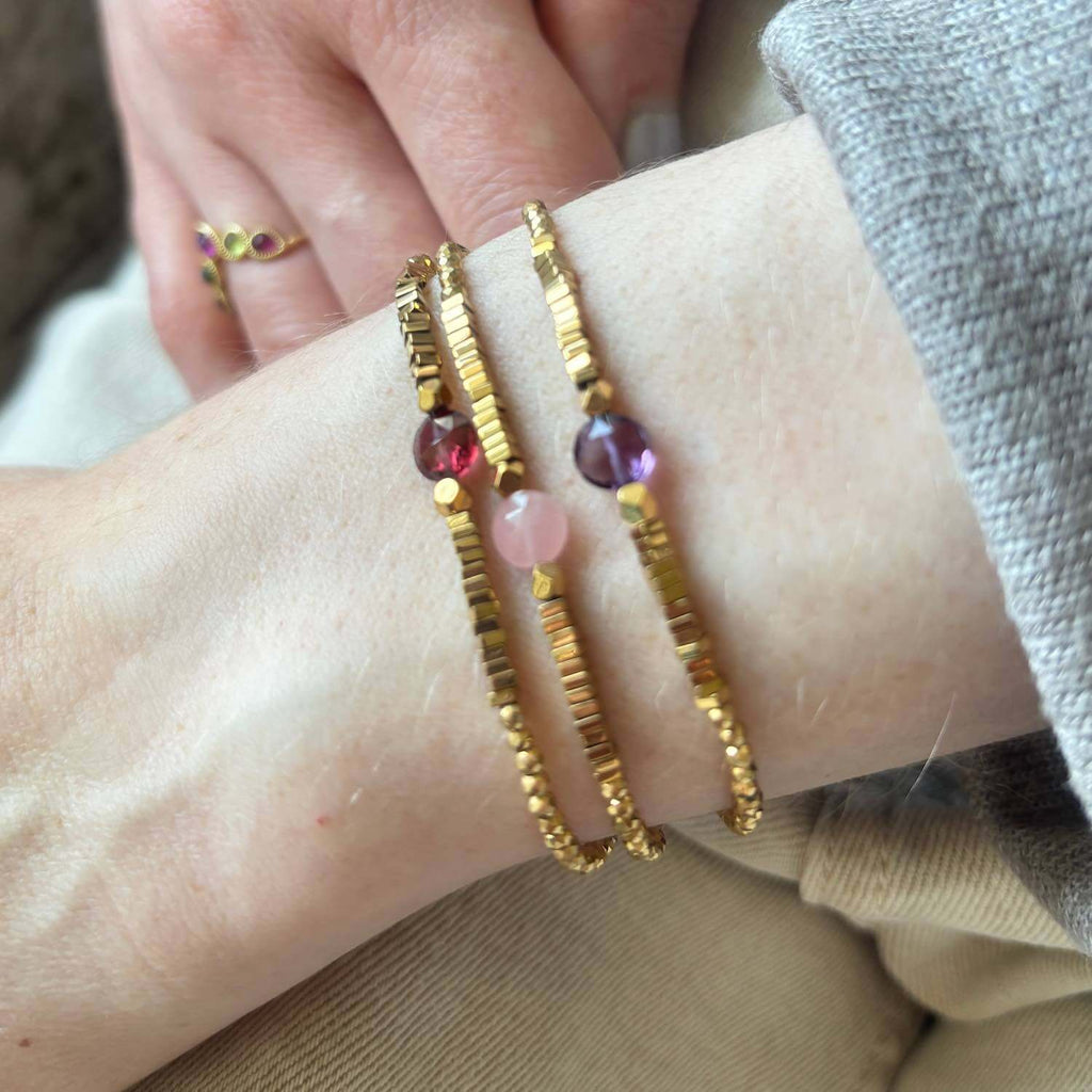 Close-up of a wrist wearing 3 elegant golden Hematite beaded bracelets with a colourful central gemstone of garnet, rose quartz and amethyst