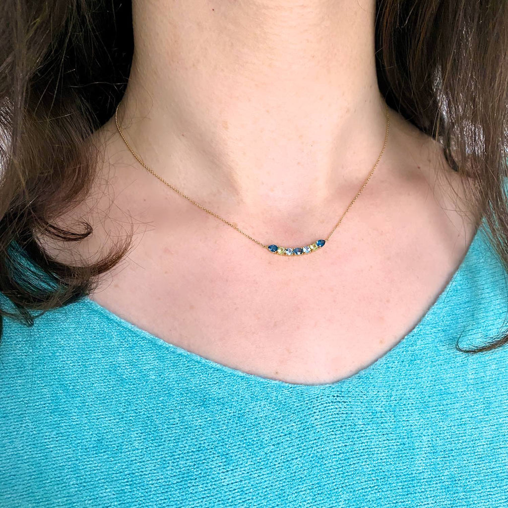 A luminous and unique 18k gold choker necklace with a central curved bar pattern of London and Swiss Blue Topaz and Peridot set on claws, displayed on a model’s neck.