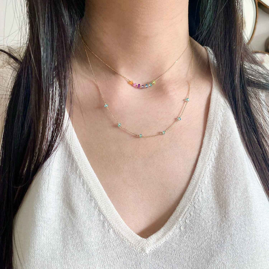 rainbow bar necklace in 18k gold with hessonite garnet, pink sapphire, amethyst, iolite, blue topaz, apatite and peridot gemstones, displayed on a model's neck layered with an apatite gemstone necklace.